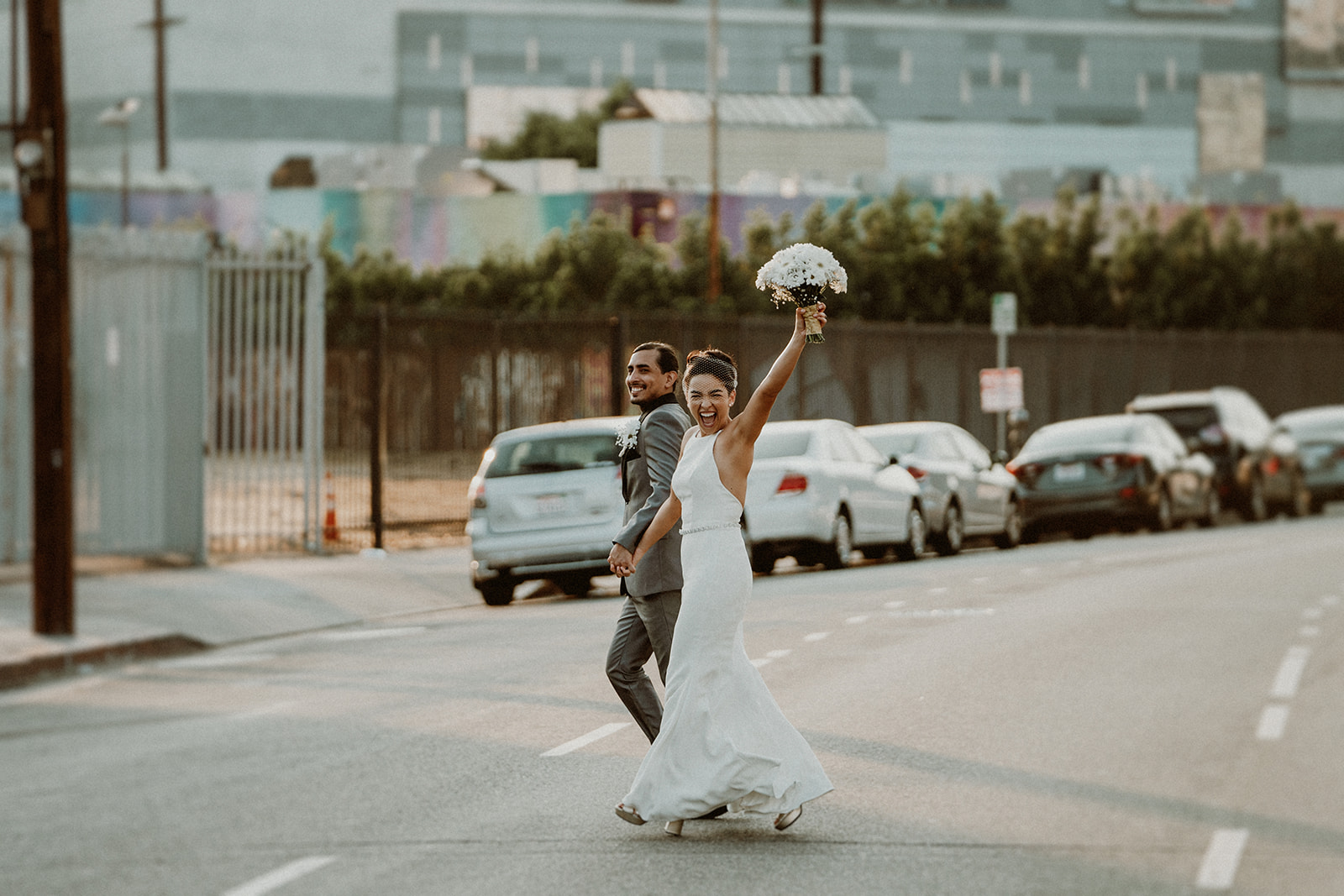 A bride and groom crossing a Southern California street, bride raising her wedding bouquet victoriously.