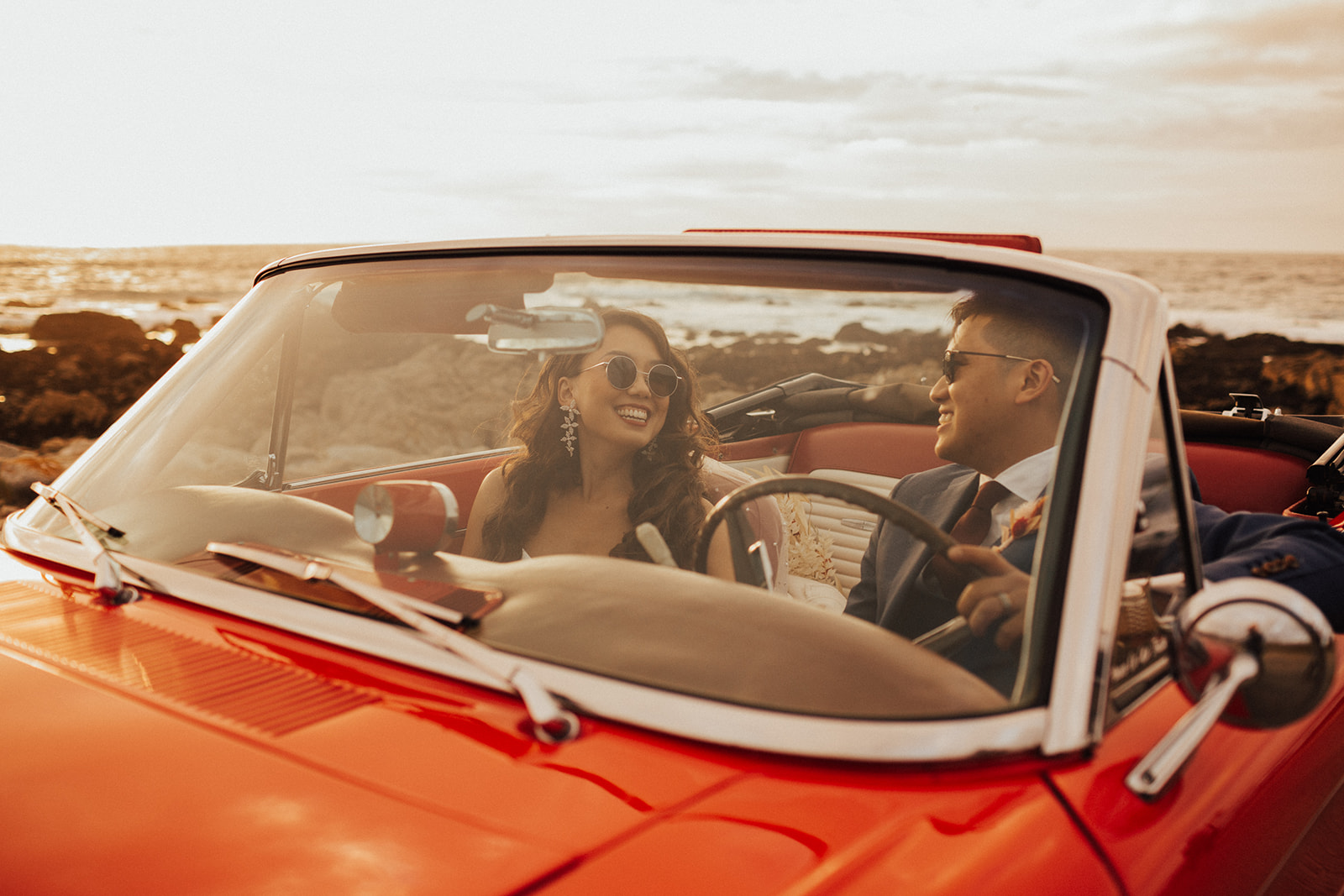 A bride and groom on the coast in a red convertible, smiling in the sunshine.